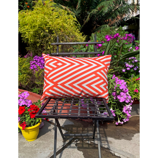 A horizontal pillow style cushion cover with red and white geometric pattern placed on a chair with a garden background.