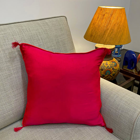 Red coloured  silk cushion cover with a subtle pink undertone, placed on a couch with a lamp and decorative objects in the background.